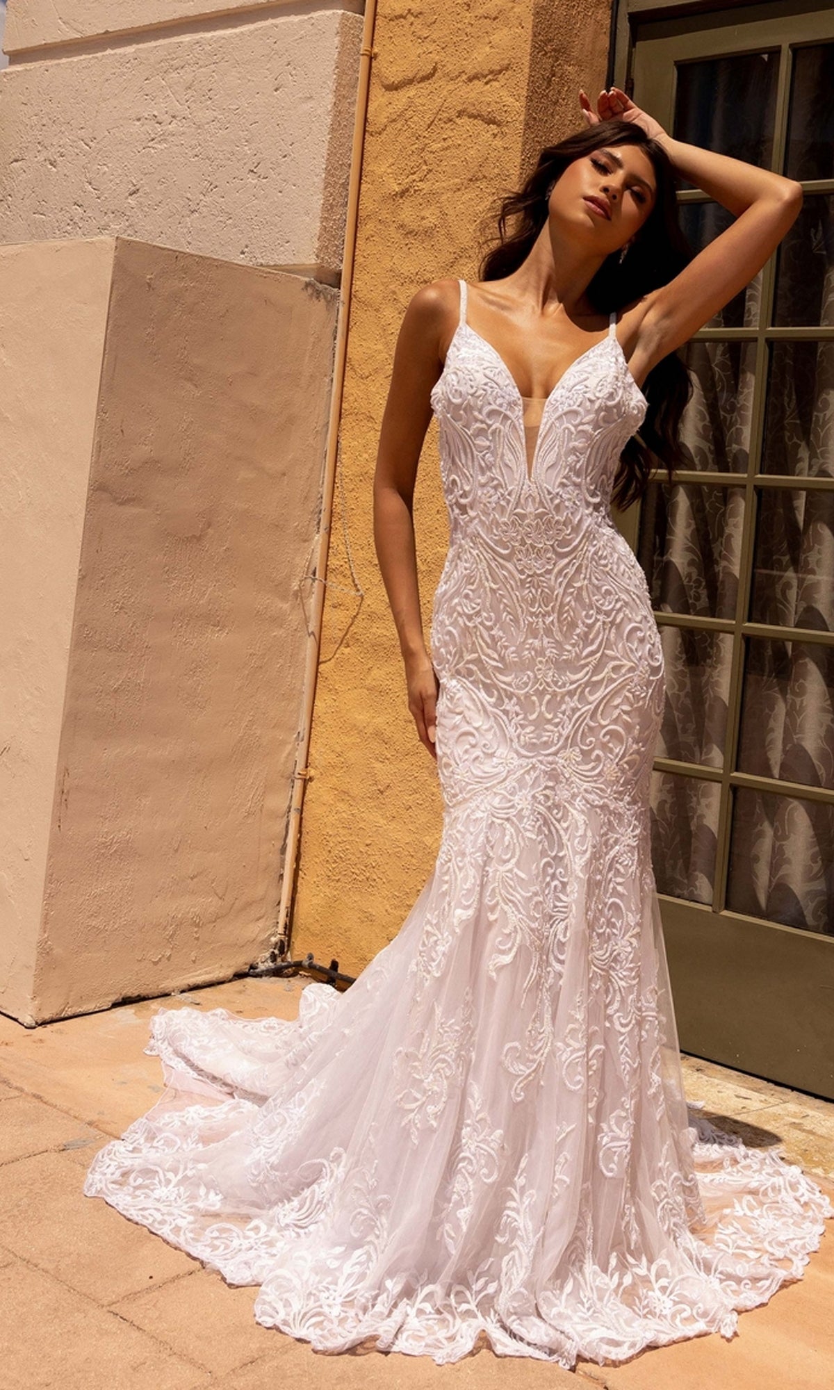 A woman stands outside by a beige wall and glass door, wearing the Long Wedding Dress 11105 by Primavera—an ivory mermaid gown with lace embroidery, thin straps, a train, and open back—her hand resting on her forehead.